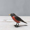 The Hand Carved Ornament - Mini Robin Bird - 3"H is a handmade wooden ornament with a red breast, dark brown head and back, yellow beak, and black legs, standing on a light wood surface against a plain gray background.