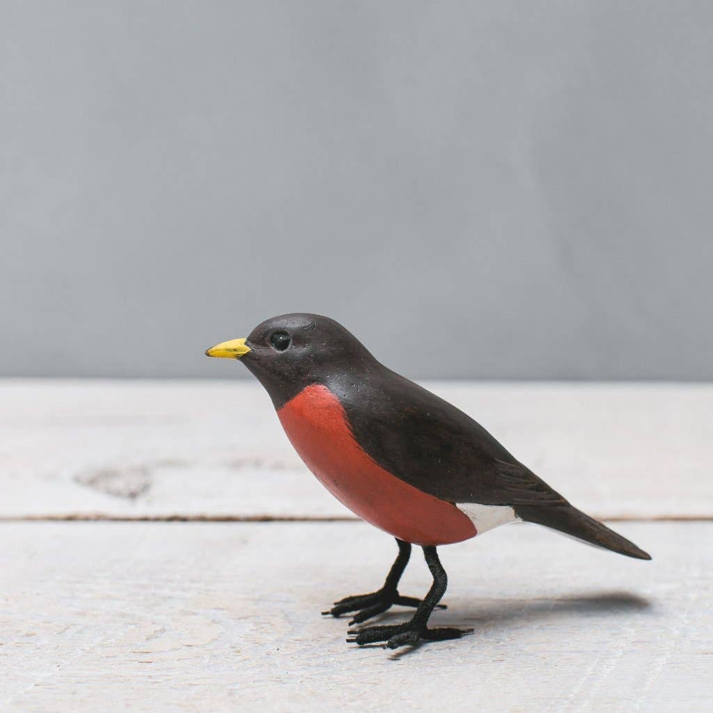 The Hand Carved Ornament - Mini Robin Bird - 3"H is a handmade wooden ornament with a red breast, dark brown head and back, yellow beak, and black legs, standing on a light wood surface against a plain gray background.
