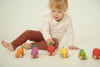 A young child in a white top and brown pants sits on the floor, reaching for the colorful Lou The Lemon Baby Car—its sensory play car design makes learning fun as they explore this wooden toy with wheels.