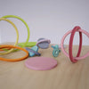 Grimm's Concentric Circles and Rings Building Set - Pastel features colorful wooden rings and disks scattered on a light wood surface, some upright, some flat or overlapping, ideal for building and balance games against a plain background.