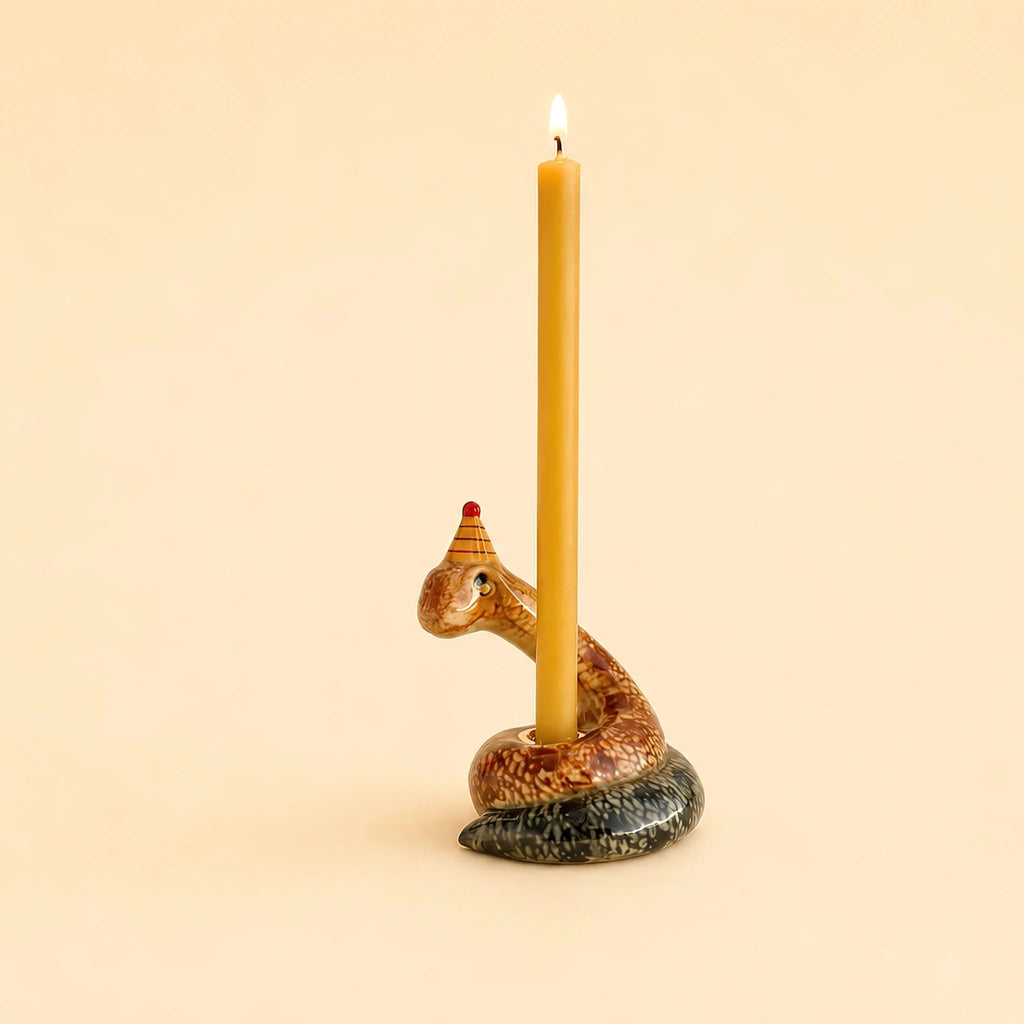 The Snake Cake Topper - Final Sale is a coiled, snake-shaped candle holder wearing a small party hat that displays a tall, lit yellow candle against a plain beige background, making it a delightful handcrafted keepsake and collectible birthday gift.