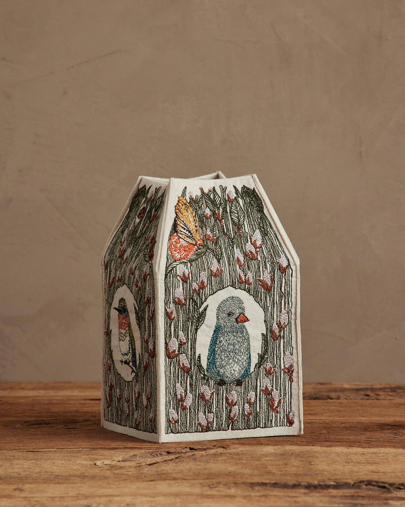 The Coral & Tusk Birdhouse Tissue Box Cover features embroidered birds and butterflies on fabric, displayed on a wooden surface against a beige wall.