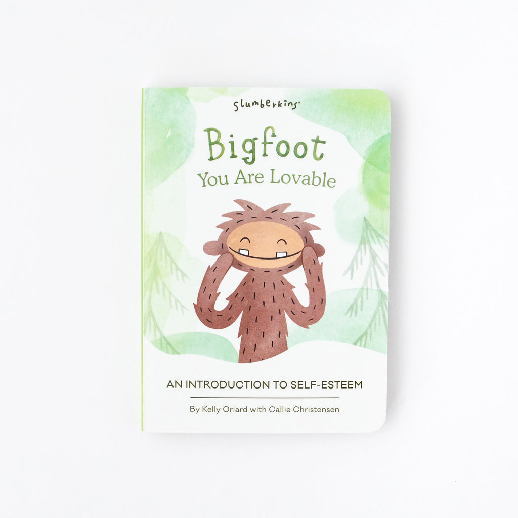 A book titled "Slumberkins Bigfoot Snuggler + Intro Book - Self Esteem" by Kelly Oriard with Callie Christensen, featuring a cartoon Bigfoot in a forest setting on the cover