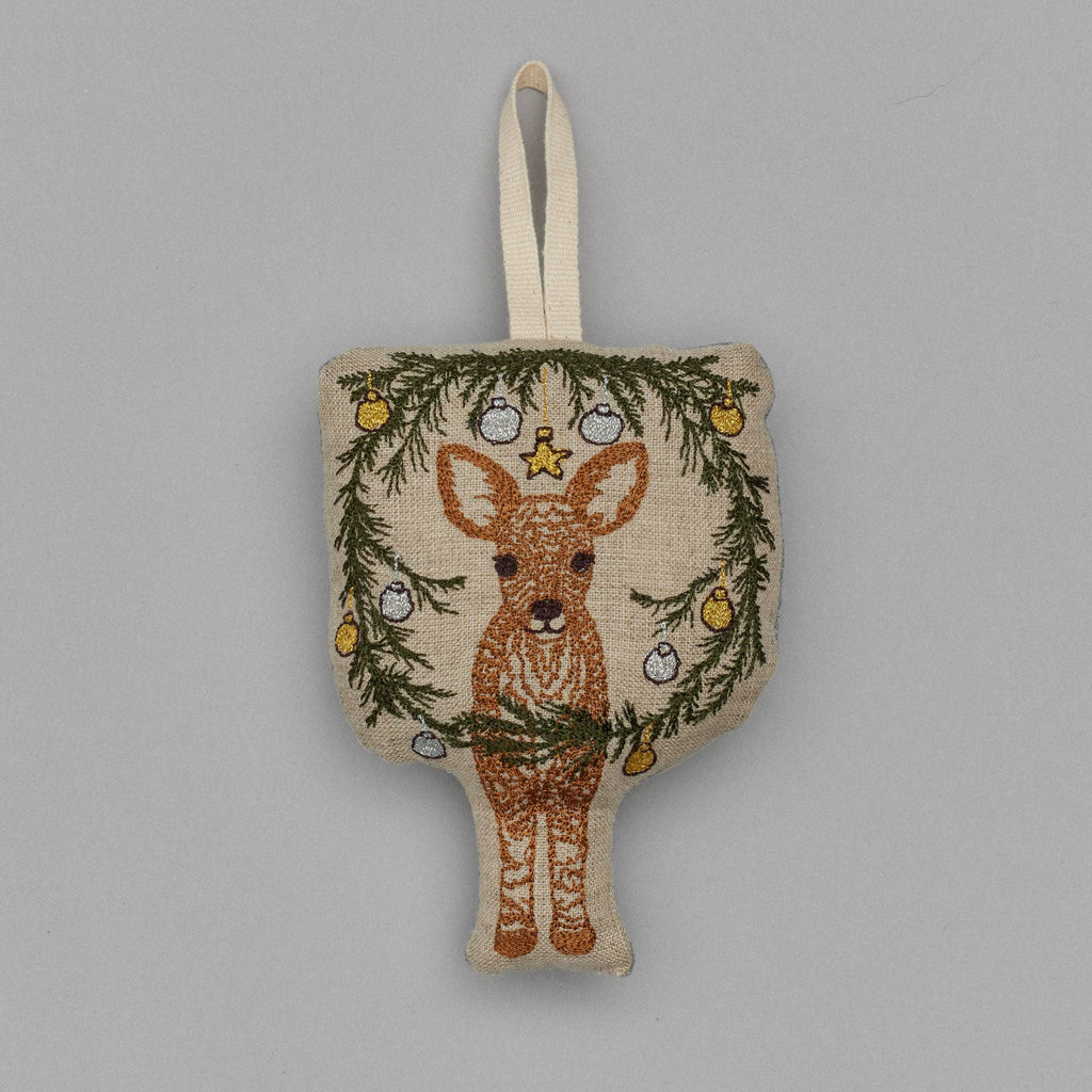 The Togetherness Deer Ornament features an embroidered deer nestled within a woodland holiday display of pine branches and baubles. It includes a loop for hanging, with a plain gray background that offers the perfect backdrop for this charming Christmas wreath.