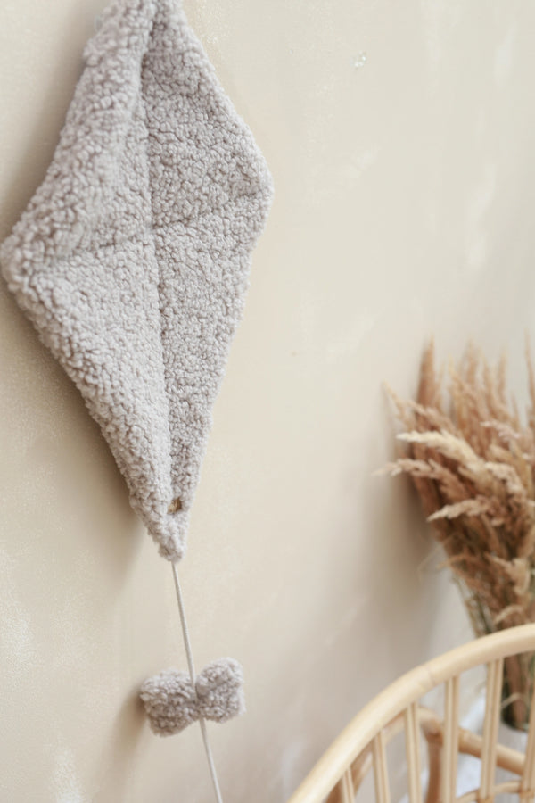 A Boucle Kite Wall Decor in taupe, made of soft teddy fabric, hangs on a beige wall, adding a charming touch to a children's room. Attached below it is a small bow of the same material on a string. In the foreground, there is a rattan chair with dried wheat stalks arranged in a nearby vase.