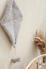 A Boucle Kite Wall Decor in taupe, made of soft teddy fabric, hangs on a beige wall, adding a charming touch to a children's room. Attached below it is a small bow of the same material on a string. In the foreground, there is a rattan chair with dried wheat stalks arranged in a nearby vase.