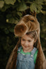A young child in denim overalls grins while wearing the Costume - Brown Horse head outdoors, standing before lush green foliage.