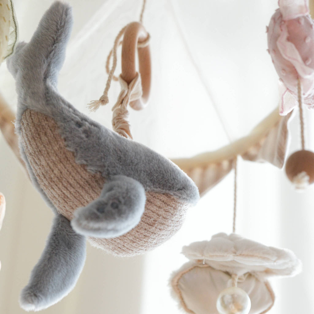 A close-up of the Nautical Dreams Mobile shows a plush whale and other soft, sea-inspired shapes with wooden beads in soothing, neutral colors—perfect for creating a calming nursery atmosphere.