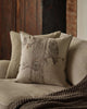 A Coral & Tusk Owl Family Tree Pillow, embroidered with three owls on a tree, rests on a beige couch with a brown knitted throw, set before rustic wooden and brick walls. The natural flax pillow adds charm to the cozy space.
