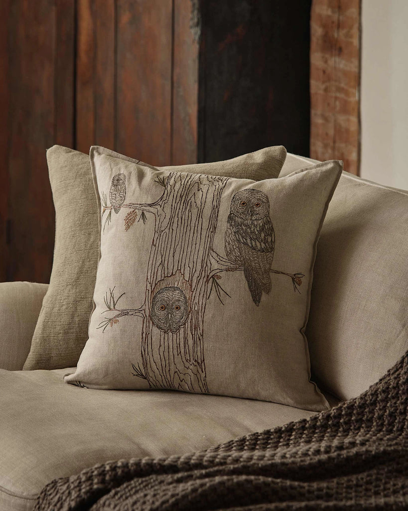 A Coral & Tusk Owl Family Tree Pillow, embroidered with three owls on a tree, rests on a beige couch with a brown knitted throw, set before rustic wooden and brick walls. The natural flax pillow adds charm to the cozy space.