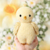 A hand holds the Cuddle + Kind Baby Bouclé Chick (Buttercup), a soft, plush yellow collectible toy with small wings, black eyes, and a tiny beak, sitting against a blurred background of white and yellow flowers.