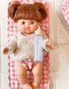 A baby doll with brown hair styled in pigtails rests on a pink and white checkered blanket, dressed in a beige sweater and pink diapers. Part of the "Tiny Tummies Pink Gift Box - The Essentials" by Tiny Harlow, it holds a clear baby bottle with a pink cap, making it an ideal addition to your collection of doll essentials.