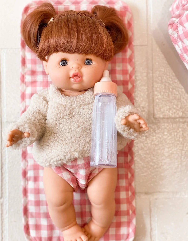 A baby doll with brown hair styled in pigtails rests on a pink and white checkered blanket, dressed in a beige sweater and pink diapers. Part of the "Tiny Tummies Pink Gift Box - The Essentials" by Tiny Harlow, it holds a clear baby bottle with a pink cap, making it an ideal addition to your collection of doll essentials.