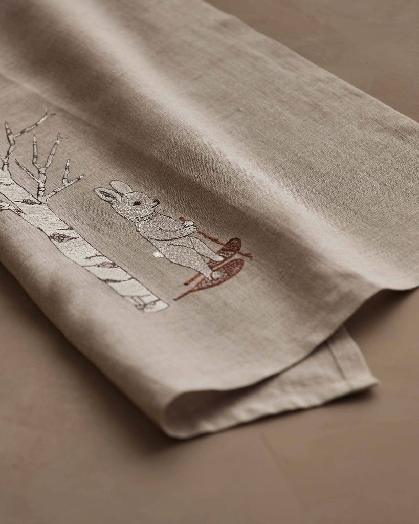 The Coral & Tusk Snowshoe Hare Tea Towel features an embroidered hare in a scarf beside a white aspen, with a red bird perched on the scarf—an ideal accent to add winter charm to your décor.