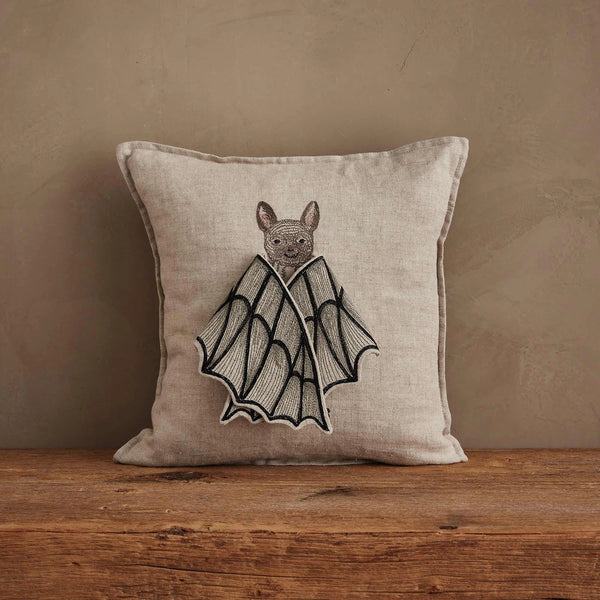 The Coral & Tusk Bat Wing Surprise Pillow—a beige throw pillow featuring an embroidered bat with wings folded like a butterfly—rests on a wooden surface against a textured beige wall.