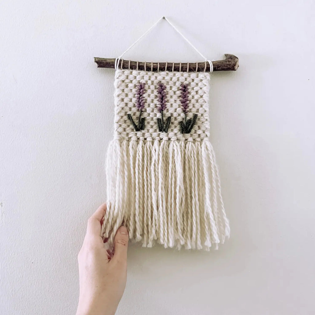 Hand holding a Handmade Lavender Wall Hanging, woven with white threads onto a driftwood stick, against a plain white wall.