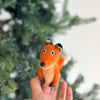 In front of a blurred green Christmas tree, a hand displays the Fox Finger Puppet, a small felted toy featuring blue eyes, ideal for storytelling.