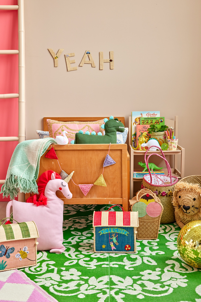 A colorful children’s room features a crib, plush unicorn pillow, patterned boxes, a dragon toy, books, a wicker bear basket, and a green patterned rug. Next to the crib is the charming Raffia Kids Bag with Circus Theme, sustainably handmade from raffia. The word “YEAH” decorates the wall above the crib.
