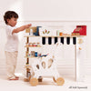 A small child plays pretend shopkeeper at a Le Toy Van wooden market stand with shelves, using the Cash Register & Money set, miniature groceries, and a white toy shopping cart in a bright, minimalist room.