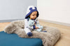 A baby in a hooded onesie with bear ears sits on a fluffy gray rug, cuddling the My First Steiff Blue Teddy Bear, 10 Inches, in a cozy room with wooden floors and white walls.