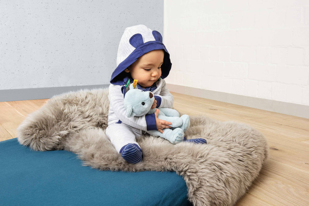 A baby in a hooded onesie with bear ears sits on a fluffy gray rug, cuddling the My First Steiff Blue Teddy Bear, 10 Inches, in a cozy room with wooden floors and white walls.