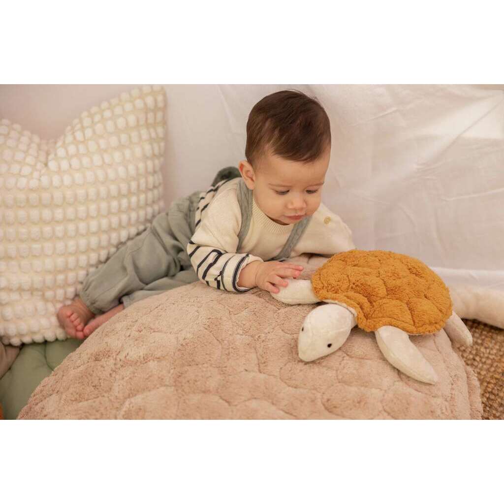 A baby in grey overalls and a striped shirt lies on a textured beige cushion, reaching for the Senger Naturwelt Cuddly Animal - Turtle made from organic cotton. A cream knitted pillow appears in the background.