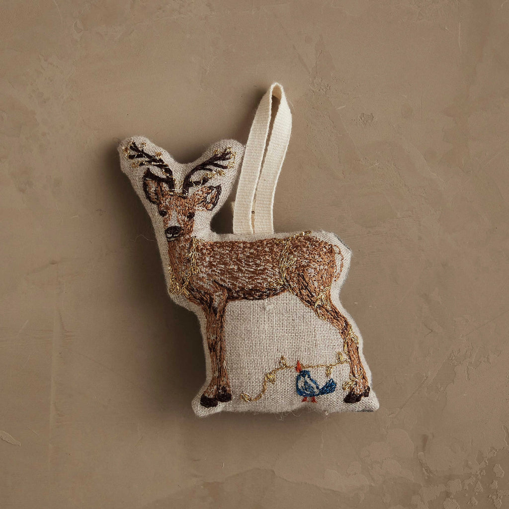 The Coral & Tusk Deer with Lights Ornament is a woodland-inspired deer ornament with embroidered details and a blue bird perched on string lights, hanging by a loop against a beige background—perfect for your Christmas tree.