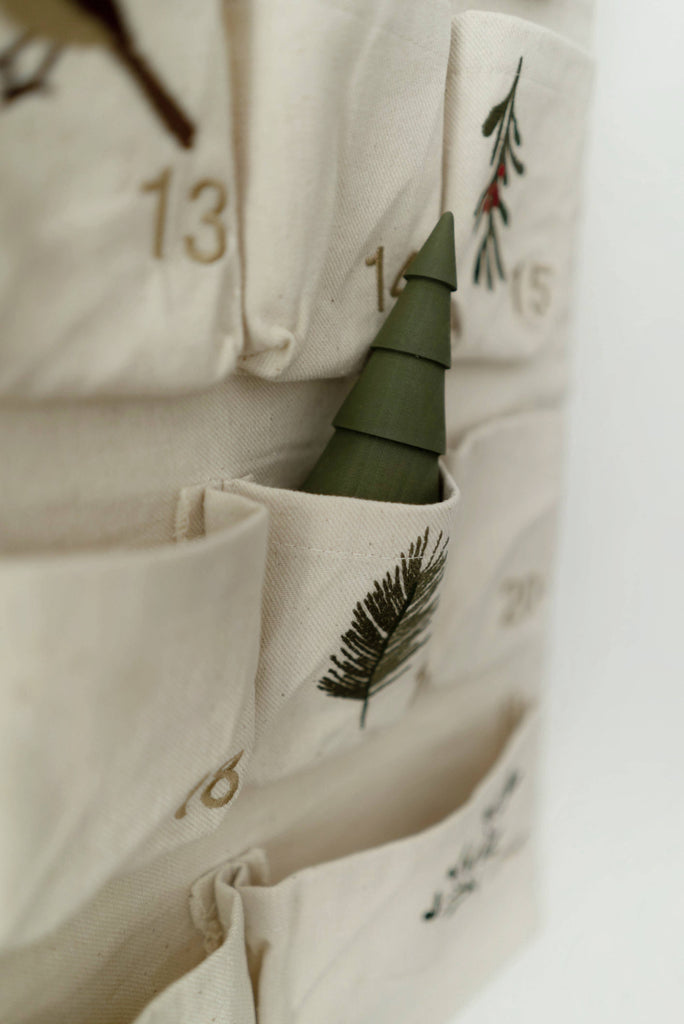A close-up of the Sabo Concept Christmas Cotton Advent Calendar, featuring beige cotton with numbered pockets. One pocket contains a small green tree, and some pockets are accented with embroidered branches.