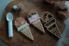 The Ice Cream Cutter Set - Eco Playdough Tool features three decorated ice cream cone cookies, a plastic spoon, an eco-friendly playdough tool, cookie cutters, rainbow sprinkles, and dough arranged on a wooden board—perfect for creative sensory play.