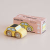 A small yellow wooden toy car sits in front of its pink and white box labeled “No. 10 GT60 Fifi | Made in England” and “Limited Edition.” This classic miniature features charming illustrations on the box that perfectly complement the vintage design of the delightful toy car.