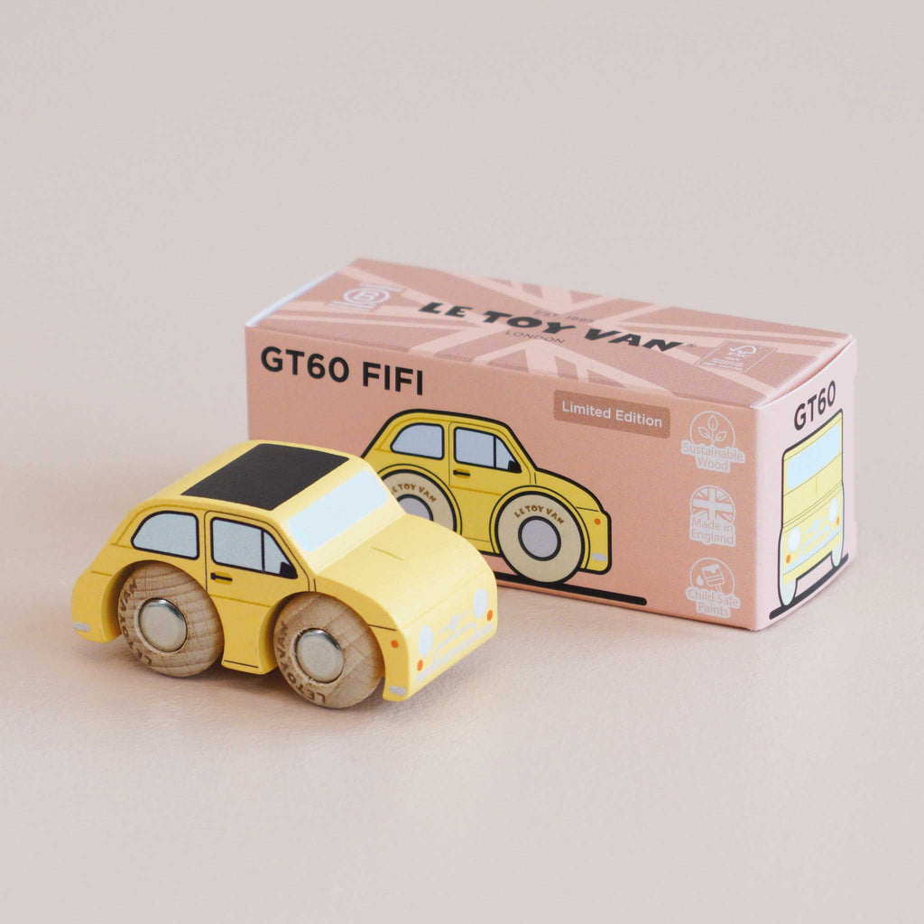 A small yellow wooden toy car sits in front of its pink and white box labeled “No. 10 GT60 Fifi | Made in England” and “Limited Edition.” This classic miniature features charming illustrations on the box that perfectly complement the vintage design of the delightful toy car.