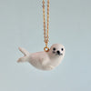 A small, white Seal Necklace hangs from a delicate 24k gold plated chain against a light blue background. The hand painted jewelry piece has black eyes and whiskers, giving it a cute and whimsical appearance.