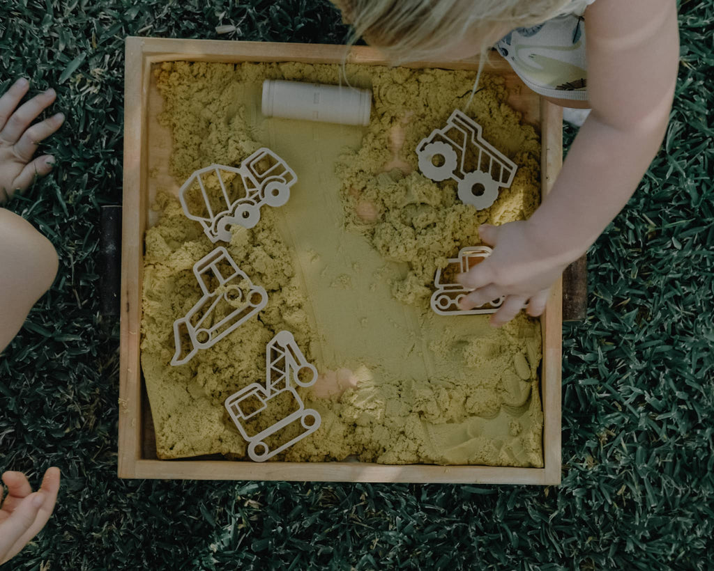 A child engages in sensory play with construction vehicle cutouts from the Construction Cutter Set - Eco Playdough Tool in a wooden box filled with yellow sand, while another child's hand is visible nearby on the green grass.