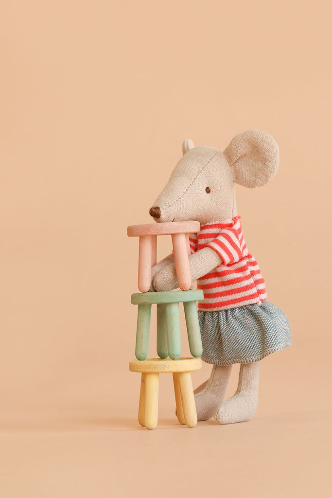 A stuffed mouse in a red striped shirt and blue skirt stands upright, stacking the Maileg Stool Set, Mouse – Light Rose/Light Mint, featuring small pastel-colored stools crafted from beech wood with a charming vintage paint finish, against a plain beige background.