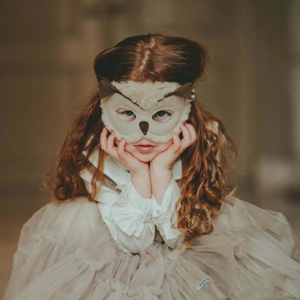 A young girl with curly hair wearing a Donsje Tieri Mask | Owl on her forehead and dressed in a frilly white dress, holding her cheeks with both hands, looking directly at the