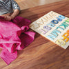 A child wearing a striped shirt sits at a wooden table, partially covered by a bright pink cloth. On the table is Grimm's ABC Game, a wooden puzzle board featuring colorful letters of the alphabet, all made from non-toxic materials.