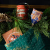 A green woven stocking holds “the poet’s garden” book, a Pollinator Seed Pop (Single) with a floral pattern, and a red “Rockin’ Around” tin, all arranged among evergreen branches and festive decorations.
