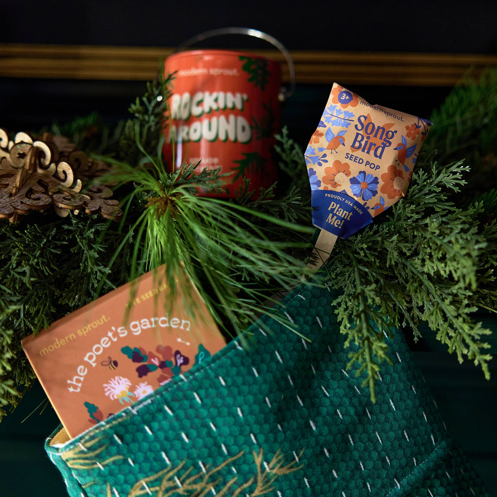 A green woven stocking holds “the poet’s garden” book, a Pollinator Seed Pop (Single) with a floral pattern, and a red “Rockin’ Around” tin, all arranged among evergreen branches and festive decorations.