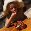 A young child in a wide-brimmed hat enjoys a strawberry at a wooden table, with a bowl of fresh strawberries in their non-toxic Pepa the Apple Bowl & Spoon Set. Sunlight casts warm light and shadows over the inviting scene.