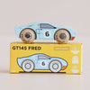 A classic wooden racing car painted in blue with the number 26 is placed on a yellow box labeled “GT145 Fred | Made in England.” The box features icons for sustainable wood, made in England, and child-safe paints.