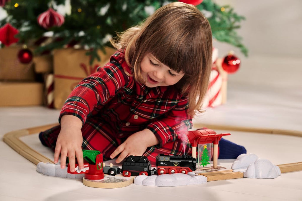 A young child in a red plaid dress plays with the BRIO World Christmas Steaming Train Set on the floor near a decorated Christmas tree and wrapped presents.