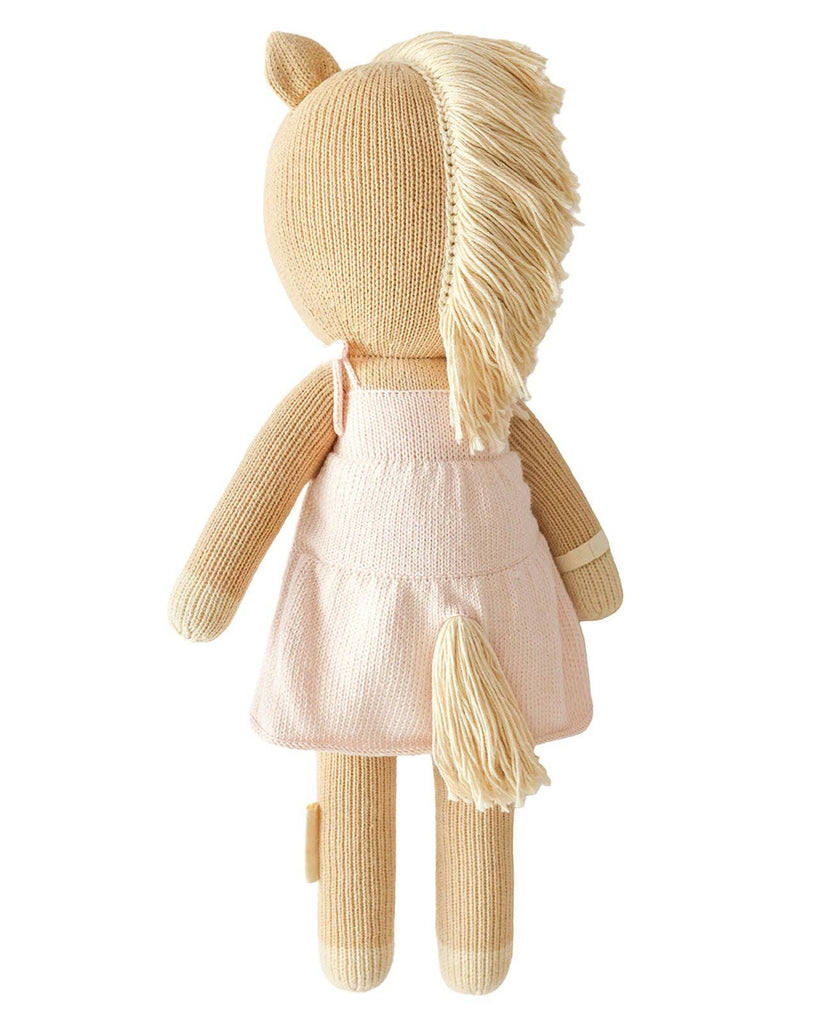 Cuddle + Kind Kelsie the Pony is a hand-knit, fair trade plush toy featuring light brown yarn skin, cream hooves, cream mane and tail, and a light pink dress. The doll is shown from the back.