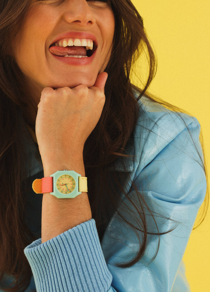 A smiling woman in a blue jacket rests her chin on her hand, showing a colorful Mini Kyomo Watch - Bubble Gum, against a bright yellow background.