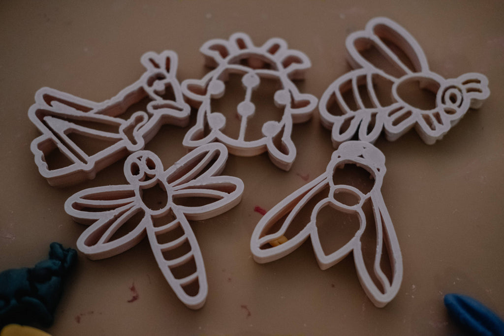 The Insect Cutter Set - Eco Playdough Tool features five insect-shaped cutters—a bee, ladybug, dragonfly, butterfly, and another winged bug—neatly arranged on a tan surface. Ideal for an eco-friendly playdough tools collection or sensory play set, these cutters inspire creativity; blue and yellow clay pieces peek out from the corners of the scene.