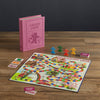 The Board Game: Candy Land Vintage Bookshelf Edition is displayed on a wooden table with colorful pieces, cards, and a bright winding board. The pink game box stands upright in the background, designed to look like a hardcover book.