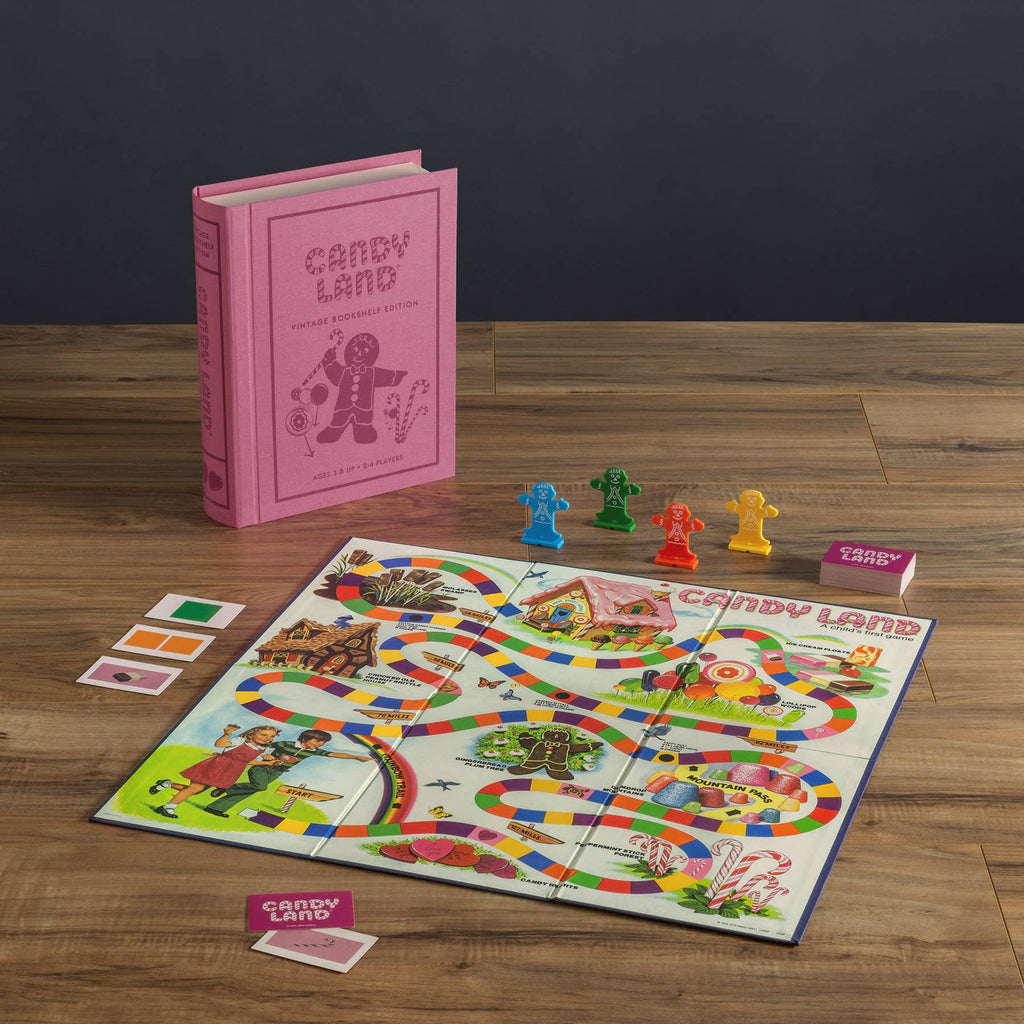 The Board Game: Candy Land Vintage Bookshelf Edition is displayed on a wooden table with colorful pieces, cards, and a bright winding board. The pink game box stands upright in the background, designed to look like a hardcover book.