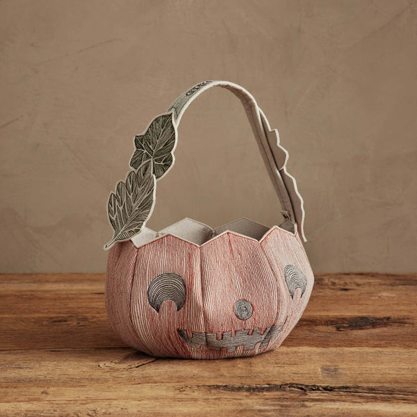The Coral & Tusk Jack-o'-Lantern Basket, featuring a stitched smiling face and leaf-patterned handle, sits on a wooden surface with a neutral background.