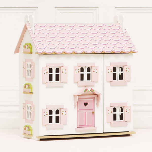 Sophie's Wooden Dolls House features a pastel pink finish, scalloped roof, heart-shaped door, pink shutters, and window flower boxes. This charming wooden dolls house is pictured on a plain white background.