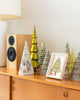 A wooden cabinet displays cone-shaped ornaments, green and yellow glass Christmas trees, the Coral & Tusk Peek a Tree Card in a frame, a keepsake wooden speaker, and a lamp in the background.