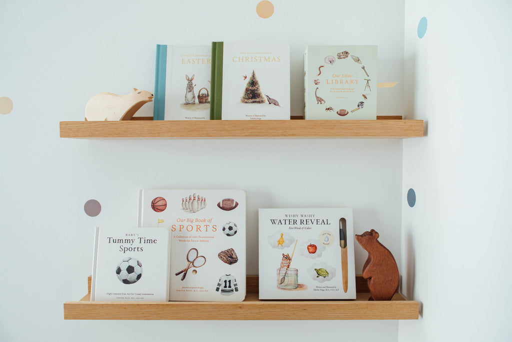 Two wooden shelves hold seven children's books and wooden animal figures. Among the books is "Baby's Tummy Time Sports," a high-contrast board book ideal for visual stimulation during tummy time. The collection features illustrations of sports, holidays, and animals. The background is a white wall decorated with pastel dot stickers.